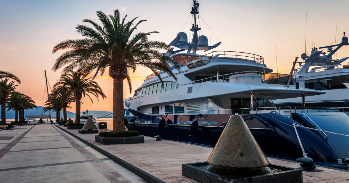 Sunset at a marina with luxury yachts, palm trees, and a paved promenade.