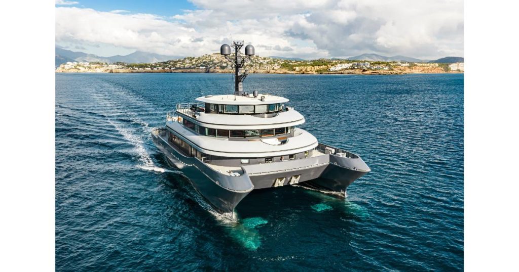 A large, modern multi-deck motor yacht cruising in deep blue water with a coastal town in the background.