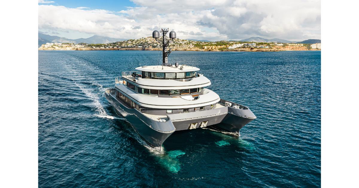 A large, modern multi-deck motor yacht cruising in deep blue water with a coastal town in the background.