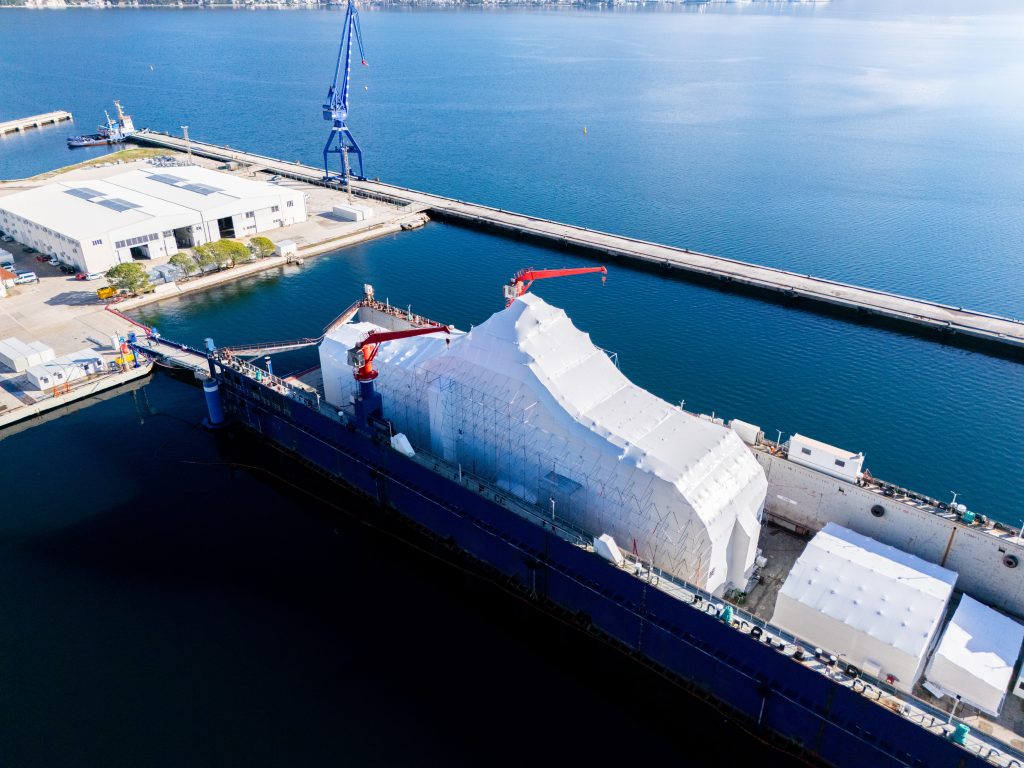Aerial view of a harbor with a tall blue crane, white warehouse buildings, and a docked cargo ship at the pier.
