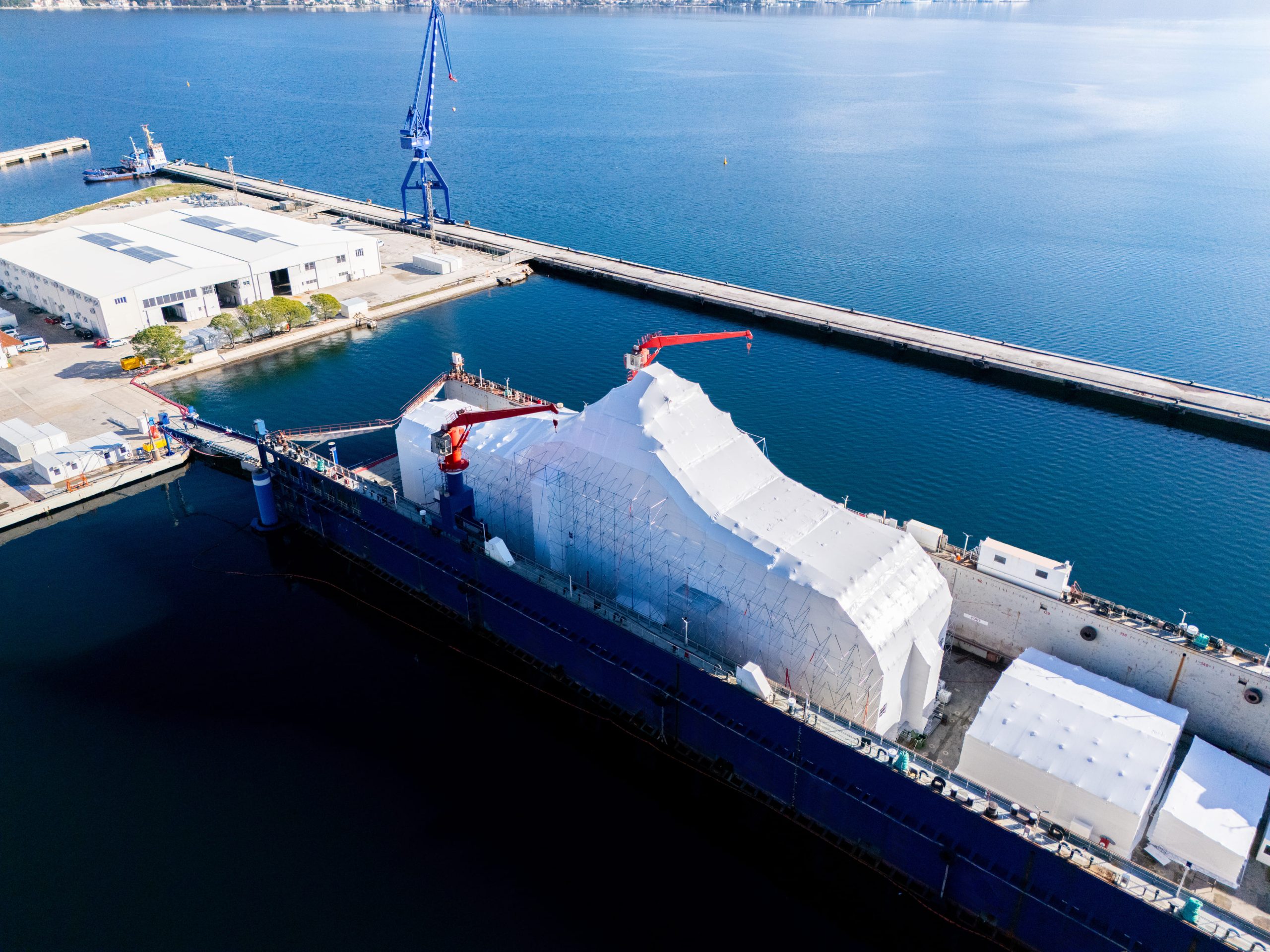 Aerial view of a harbor with a tall blue crane, white warehouse buildings, and a docked cargo ship at the pier.